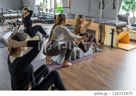 Group of women doing exercises on back spine corrector. Reformer pilates studio machine for fitness workouts in gym. Fit, healthy and strong authentical body. Fitness concept. Group of women doing exercises on back spine corrector. Reformer pilates studio machine for fitness workouts in gym. Fit, healthy and strong authentical body. Fitness concept. 126372875