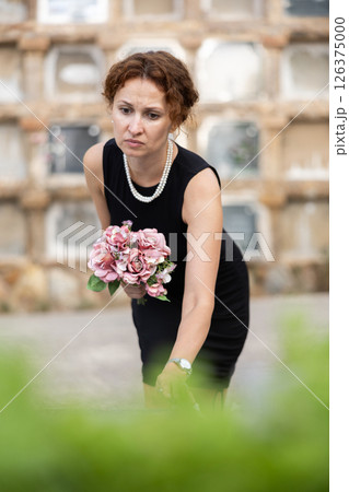 Sorrowful woman in black resting flowers on grave in cemetery 126375000