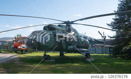 A Soviet-era helicopter rests on display in an open area, showcasing its military design and vintage aesthetic against a picturesque backdrop of greenery and buildings. 126376176