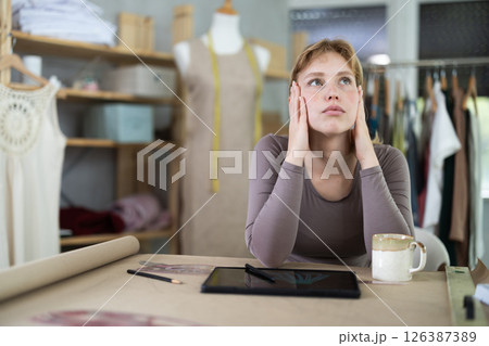 Tired female fashion designer sitting at table in sewing studio Tired female fashion designer sitting at table in sewing studio 126387389