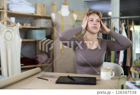 Tired female fashion designer sitting at table in sewing studio Tired female fashion designer sitting at table in sewing studio 126387538