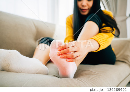 A woman seated on a sofa massages her painful foot highlighting the area of discomfort. Illustrating health care reflexology and recovery through massage and relief from pain. A woman seated on a sofa massages her painful foot highlighting the area of discomfort. Illustrating health care reflexology and recovery through massage and relief from pain. 126389492