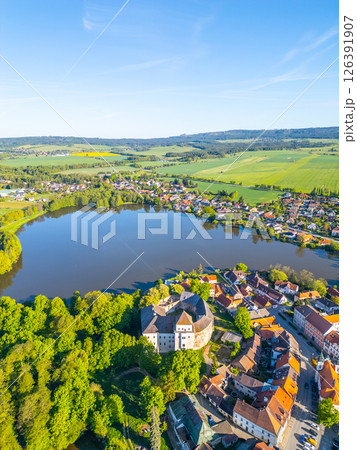 The scenic view showcases Rozmital pod Tremsinem, featuring a prominent castle and a tranquil lake. The town is nestled among lush greenery under a clear blue sky. 126391907