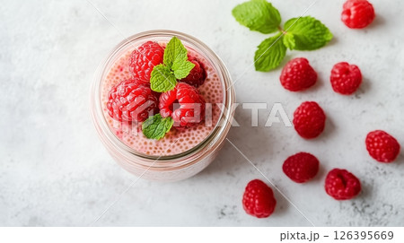 A jar of creamy chia pudding topped with fresh raspberries and mint leaves, placed on a clean white table with soft natural light, concept of healthy eating 126395669