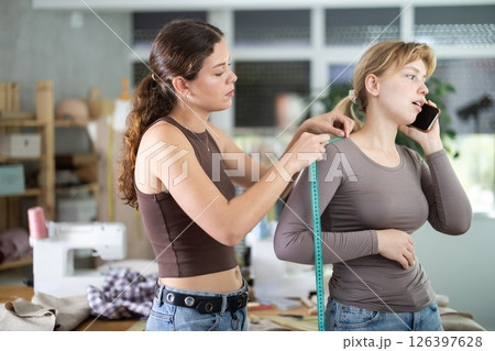 Young tailoress measuring female client speaking on mobile 126397628