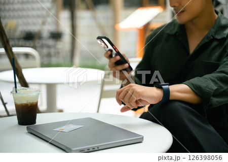 Smiling man relaxes at an outdoor cafe and using smartphone beside a closed laptop Smiling man relaxes at an outdoor cafe and using smartphone beside a closed laptop 126398056