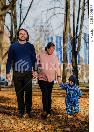 A loving couple strolls through a sunlit park with their young son, surrounded by the vibrant colors of autumn, enjoying a joyful and peaceful family moment together. 126399677