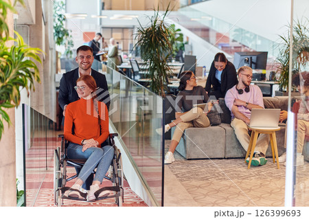 A company director assists his business colleague in a wheelchair, helping her navigate to their startup office, where they work alongside their diverse team of colleagues, emphasizing inclusivity and 126399693