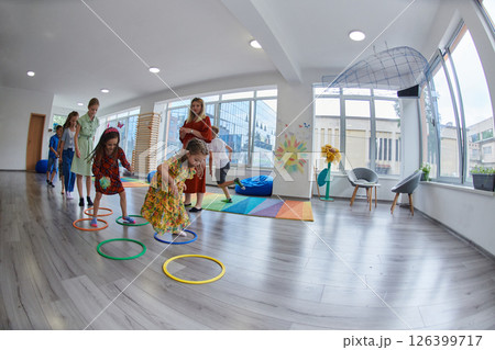Small nursery school children with female teacher on floor indoors in classroom, doing exercise. Jumping over hula hoop circles track on the floor. 126399717