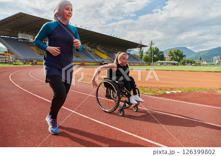A Muslim woman in a burqa running together with a woman in a wheelchair on the marathon course, preparing for future competitions. A Muslim woman in a burqa running together with a woman in a wheelchair on the marathon course, preparing for future competitions. 126399802