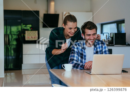 A young married couple is talking to parents, family and friends on a video call via a laptop while sitting in the living room of their modern house. A young married couple is talking to parents, family and friends on a video call via a laptop while sitting in the living room of their modern house. 126399876