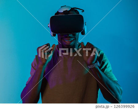 African American man immerses himself in a thrilling horror gaming experience using VR glasses, creating an isolated and intense atmosphere against a striking blue background African American man immerses himself in a thrilling horror gaming experience using VR glasses, creating an isolated and intense atmosphere against a striking blue background 126399926