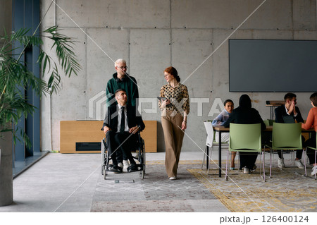 A diverse team of business colleagues, including a director in a wheelchair, engages in a collaborative discussion as they move together through a modern office space 126400124