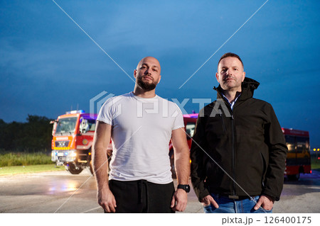 Group of firefighters, dressed in civilian clothing, stand in front of fire trucks during the night, showcasing a moment of camaraderie and unity among the team as they reflect on their duties and the 126400175