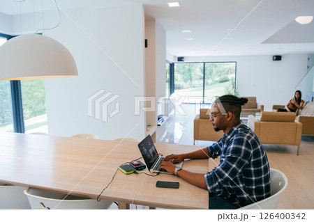 African American influencer in glasses sitting at a table in a modern living room, using a laptop for business video chat, conversation with friends and entertainment. African American influencer in glasses sitting at a table in a modern living room, using a laptop for business video chat, conversation with friends and entertainment. 126400342