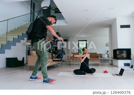 Behind the scene. Photo of a cameraman recording a woman doing yoga exercises at an online training while using and watching a laptop. 126400376