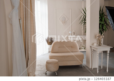 Elegant bridal room interior featuring a soft beige sofa, decorative wall accents, a white side table, and a wedding dress draped on a hanger, setting a romantic atmosphere Elegant bridal room interior featuring a soft beige sofa, decorative wall accents, a white side table, and a wedding dress draped on a hanger, setting a romantic atmosphere 126403187