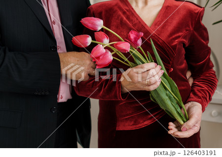 A romantic indoor scene depicting an elegant couple, the man in a suit presenting a lush bouquet of vibrant pink tulips to the woman in a red velvet dress A romantic indoor scene depicting an elegant couple, the man in a suit presenting a lush bouquet of vibrant pink tulips to the woman in a red velvet dress 126403191