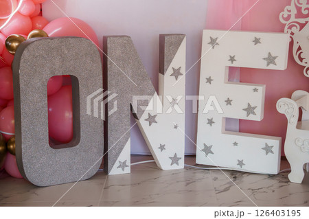 A glamorous first birthday celebration backdrop featuring large, sparkling silver "ONE" letter decorations adorned with stars, alongside a pastel pink balloon garland and a unicorn statue 126403195