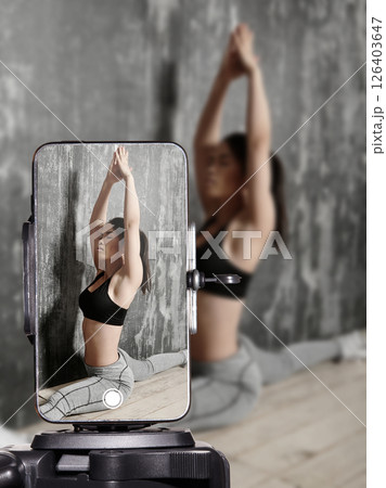 Fitness influencer doing home workout and stretching on yoga mat while live streaming on smartphone. Sporty young woman filming online fitness training for social media followers. Fitness influencer doing home workout and stretching on yoga mat while live streaming on smartphone. Sporty young woman filming online fitness training for social media followers. 126403647