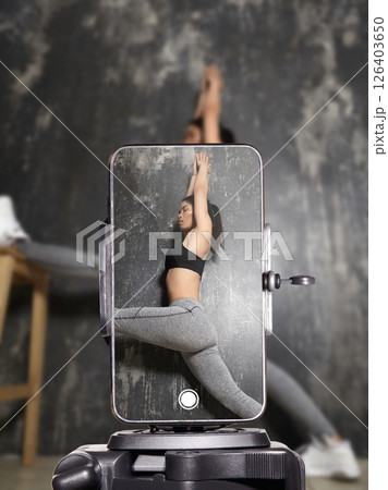 Fitness influencer doing home workout and stretching on yoga mat while live streaming on smartphone. Sporty young woman filming online fitness training for social media followers 126403650