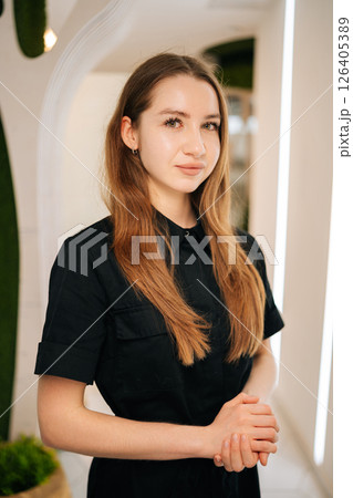 Young female therapist with long brown hair, wearing black uniform, standing in modern bright interior with white walls, green plants and soft lighting, exuding professionalism and competence. Young female therapist with long brown hair, wearing black uniform, standing in modern bright interior with white walls, green plants and soft lighting, exuding professionalism and competence. 126405389