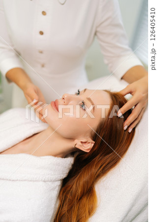 Close-up vertical shot of beautician comforting client before wellness treatment, providing caring support. Cosmetologist performing relaxing facial massage for young woman lying on massage table. 126405410