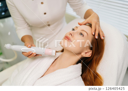 Close-up hands of unrecognizable beautician performing mesotherapy treatment, targeting facial skin rejuvenation with specialized dermatological equipment to female client in modern aesthetic clinic 126405425