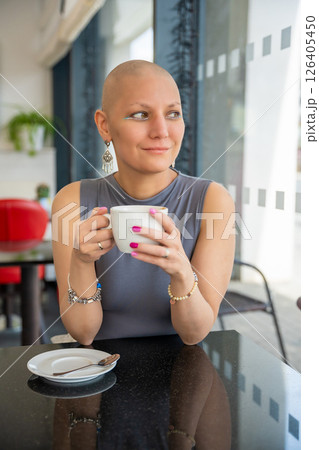 Young woman with cancer sitting in a cafe, drinking coffee and enjoying everyday life. The concept highlights that people with illnesses continue to live actively and socialize Young woman with cancer sitting in a cafe, drinking coffee and enjoying everyday life. The concept highlights that people with illnesses continue to live actively and socialize 126405450