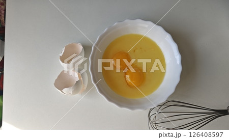 Two raw eggs in a white bowl with broken eggshells and a metal whisk on a light background. Concept of cooking preparation, natural ingredients, and simplicity in home baking 126408597