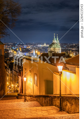 Strolling down the staircase of Prague Castle reveals a stunning view of the city bathed in golden light, with historical buildings and the skyline glistening under the night sky. 126413543