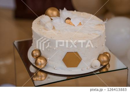 A beautifully decorated two-tier baby shower cake with a white frosting, adorned with golden balls, delicate white feathers, and a sleeping baby figurine on top A beautifully decorated two-tier baby shower cake with a white frosting, adorned with golden balls, delicate white feathers, and a sleeping baby figurine on top 126413883