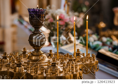 A serene close-up view of a brass candle stand adorned with lit taper candles within a richly decorated Orthodox church interior, showcasing faith A serene close-up view of a brass candle stand adorned with lit taper candles within a richly decorated Orthodox church interior, showcasing faith 126413923
