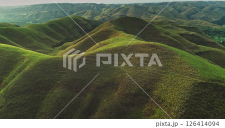 Aerial view of the undulating landscape of Sumba Island, Indonesia, bathed in the warm glow of golden hour, showcasing the beauty of nature's curves and vibrant green vegetation 126414094