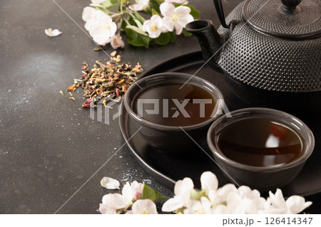 Black tea in cast iron teapot and cups decorated and white spring blossom flowers on black table. Chinese beverage. Tea ceremony. Close up. Copy space. 126414347