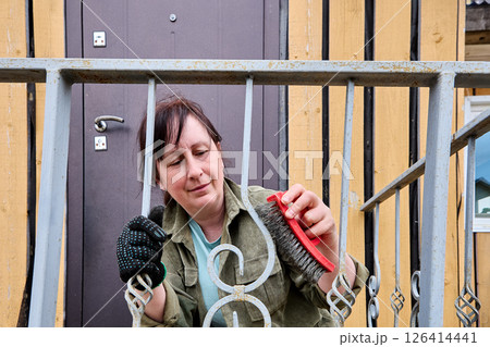 Woman using wire brush to clean rust from metal porch railing preparing surface for painting outside rustic wooden house in countryside. Woman using wire brush to clean rust from metal porch railing preparing surface for painting outside rustic wooden house in countryside. 126414441