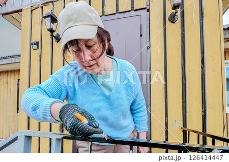 Applying black protective paint with small brush to restore metal porch railing woman in her 50s wearing cap and glove concentrates on painting process. 126414447