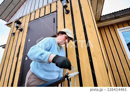 Painting metal railing with brush to protect surface from corrosion and improve appearance middle aged woman in cap works near entrance of wooden house. Painting metal railing with brush to protect surface from corrosion and improve appearance middle aged woman in cap works near entrance of wooden house. 126414461