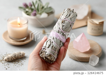 Hand holding sage smudge stick with burning candle, rose quartz crystal and herbs on soft background 126414675