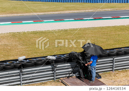 Cameraman crew stands large pro video camera on motorsport race track shaded heat black umbrella hot summer day. TV equipment safety tires fence automotive moto challenge auto racing 126416534