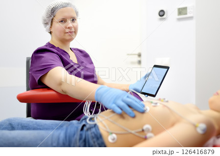 Child under ECG test with doctor. Boy patient lying in medical bed with vacuum sensors. A nurse performs an electrocardiogram of the heart in office of a medical clinic. 126416879