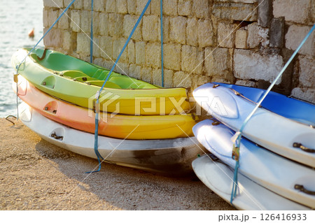 A group of kayaks stacked on top of each other lie on the shore in the sunset light. Boats ready for the tourist season on the waterfront in Perast, Montenegro A group of kayaks stacked on top of each other lie on the shore in the sunset light. Boats ready for the tourist season on the waterfront in Perast, Montenegro 126416933