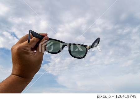 Hand holding sunglasses against cloudy sky outdoor adventure fashion nature upward view summer vibes 126419749