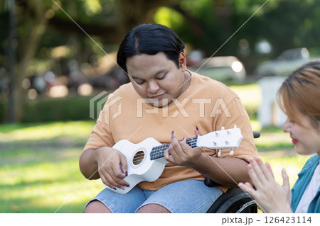 Young man in a wheelchair playing ukulele while a friend claps in appreciation. 126423114