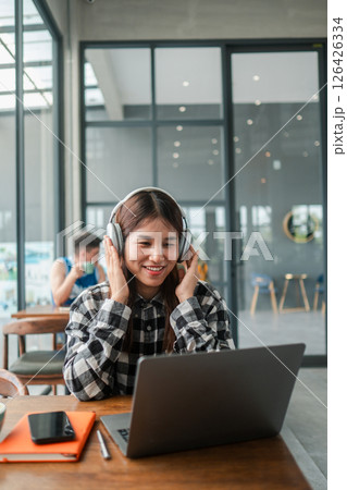 Smiling woman wearing headphones, using a laptop in a contemporary cafe environment, enjoying music and work. 126426334