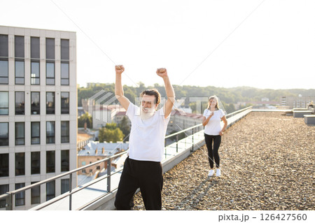 Senior man in white shirt exercising outdoors on city rooftop. Woman in background also participates in fitness activities. Scenic cityscape creates dynamic and invigorating atmosphere Senior man in white shirt exercising outdoors on city rooftop. Woman in background also participates in fitness activities. Scenic cityscape creates dynamic and invigorating atmosphere 126427560