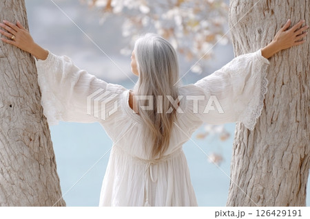 Woman in flowing white dress standing between two trees with arms outstretched, facing serene blue water in peaceful natural setting 126429191