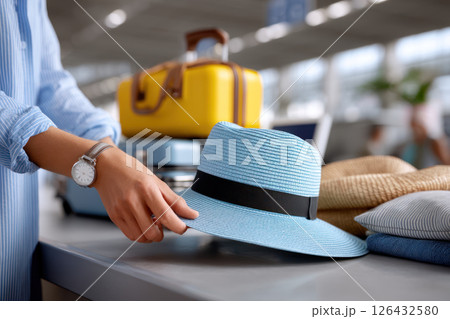 Summer traveler placing blue sunhat on carry on luggage at check in counter, preparing for vacation with stylish accessories and bright suitcase 126432580