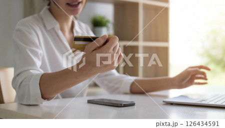 Attractive Asian woman sitting relaxed on sofa, holding credit card and smartphone probably buying something online. 126434591