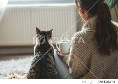 Young woman wearing a sweater freezing for winter cold with her cat. Girl is sitting over electric heater, drinking hot coffee or tea, warming up hands. Discomfort spending time at home. Coldly, chill 126436814